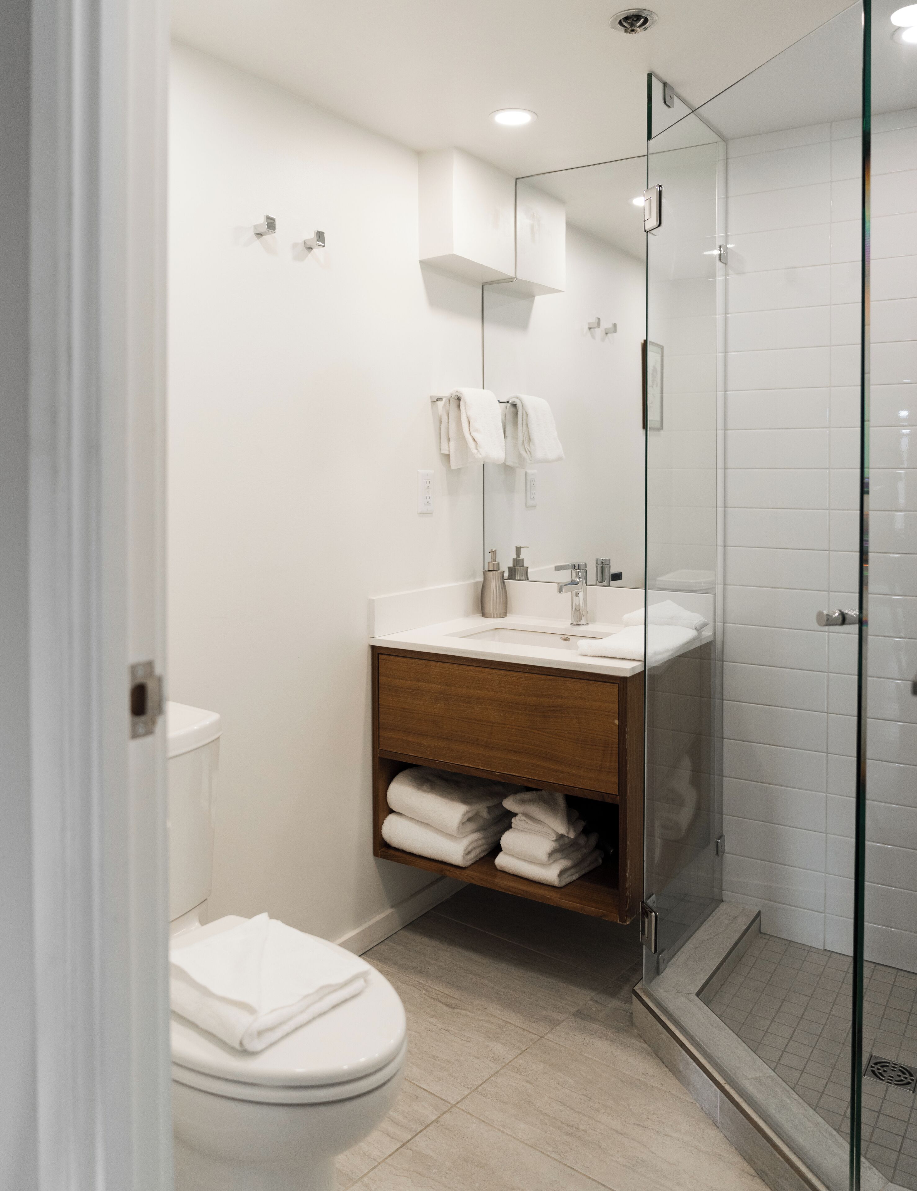 Un baño moderno con ducha envuelta en vidrio, inodoro blanco y tocador de madera con lavamanos.