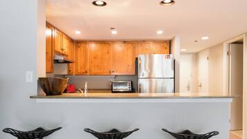 A modern kitchen with wooden cabinets, a stainless steel refrigerator, a toaster, and a bar area with stools.