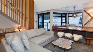 Una sala de estar moderna con un sofá modular gris, un muro de listones de madera, ventanas amplias con vistas nevadas, una mesa de comedor de madera con velas, y un telesilla al fondo.