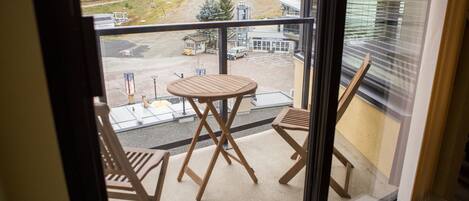A balcony with a wooden table and two chairs, overlooking a parking area with a building in the background.