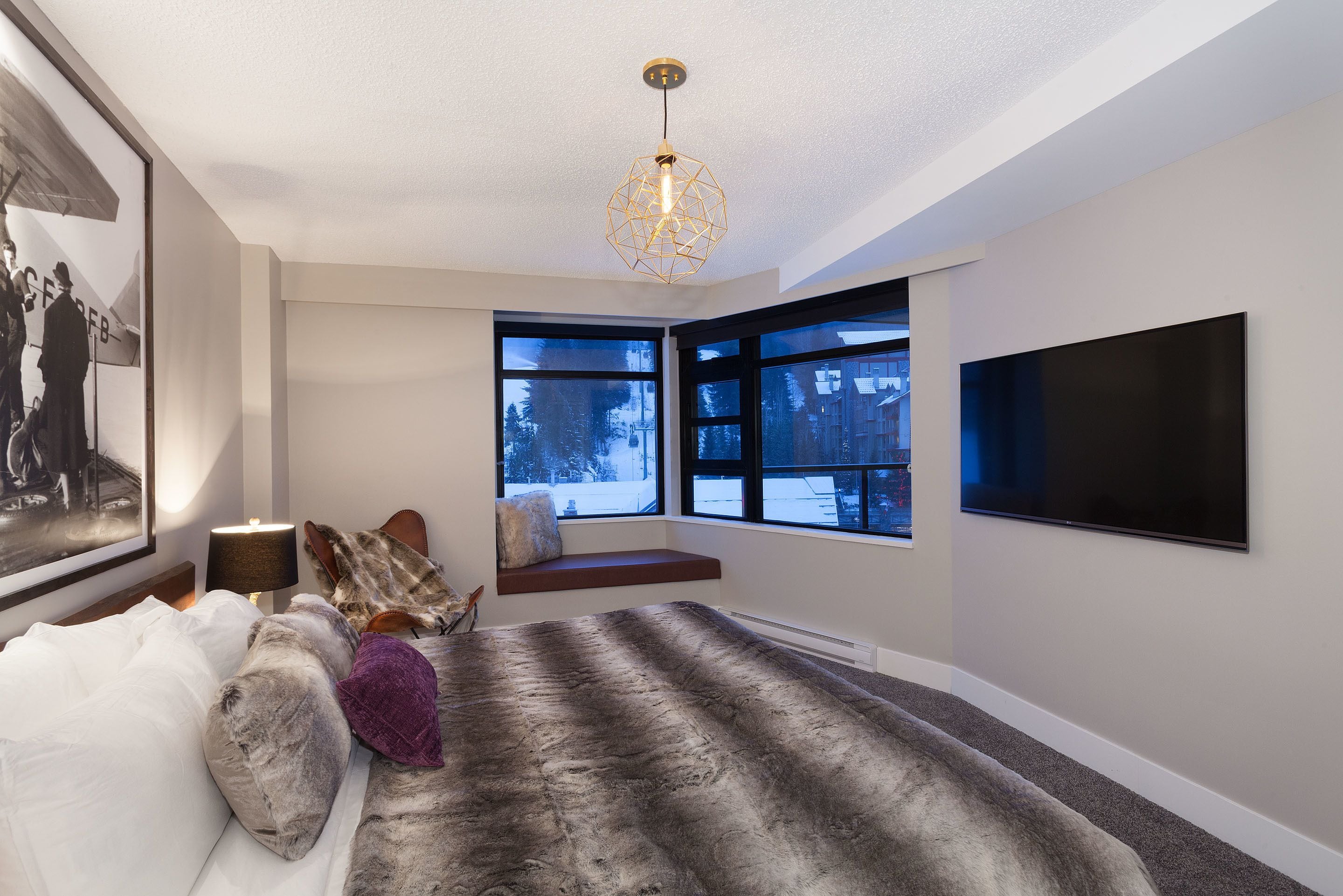 A modern bedroom with a large bed, a built-in bench, a TV, and a window with a view of trees.