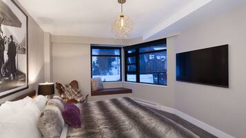 A modern bedroom with a large bed, a built-in bench, a TV, and a window with a view of trees.