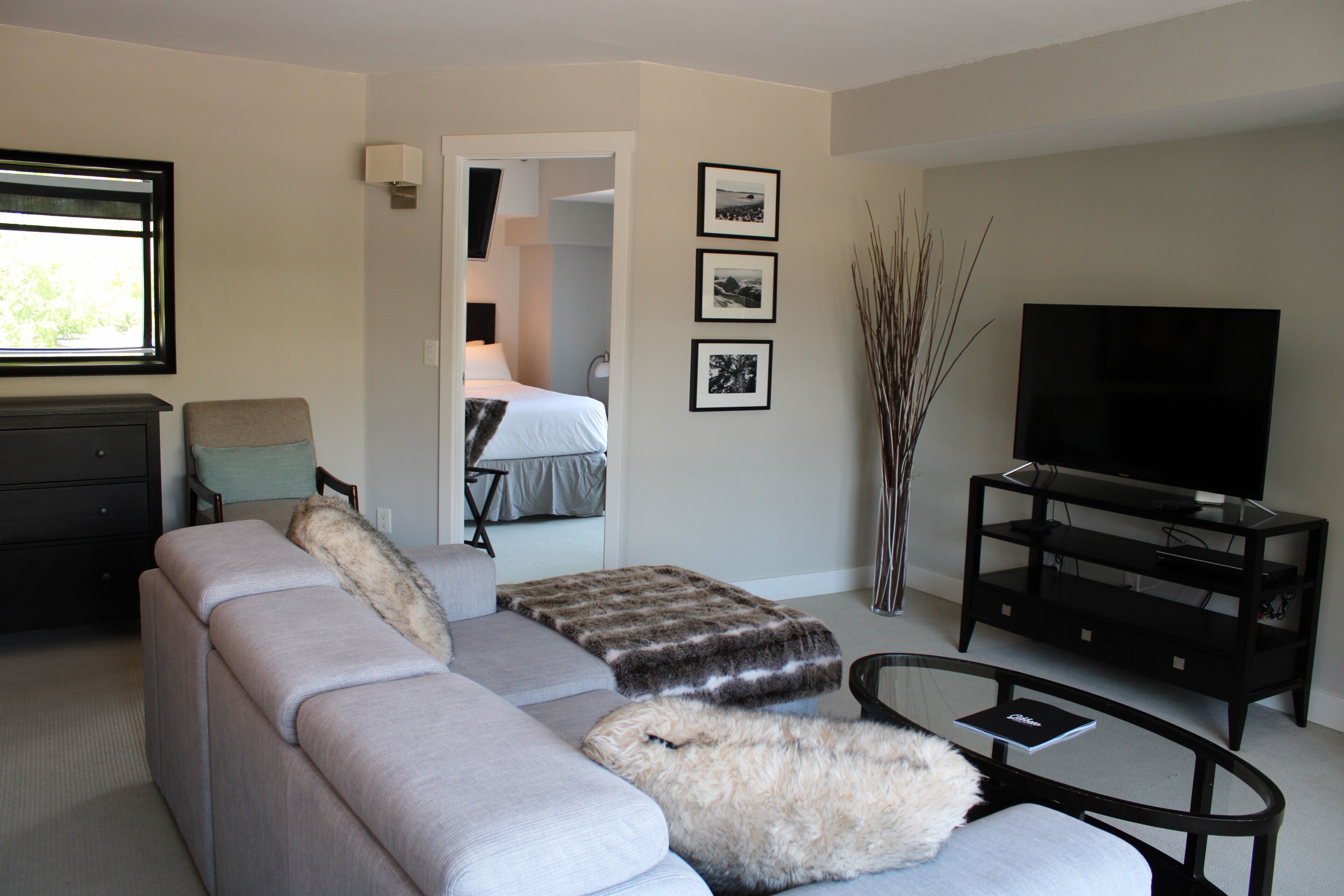 A modern living room with a grey sofa, a coffee table, a TV on a black stand, and a door leading to a bedroom.