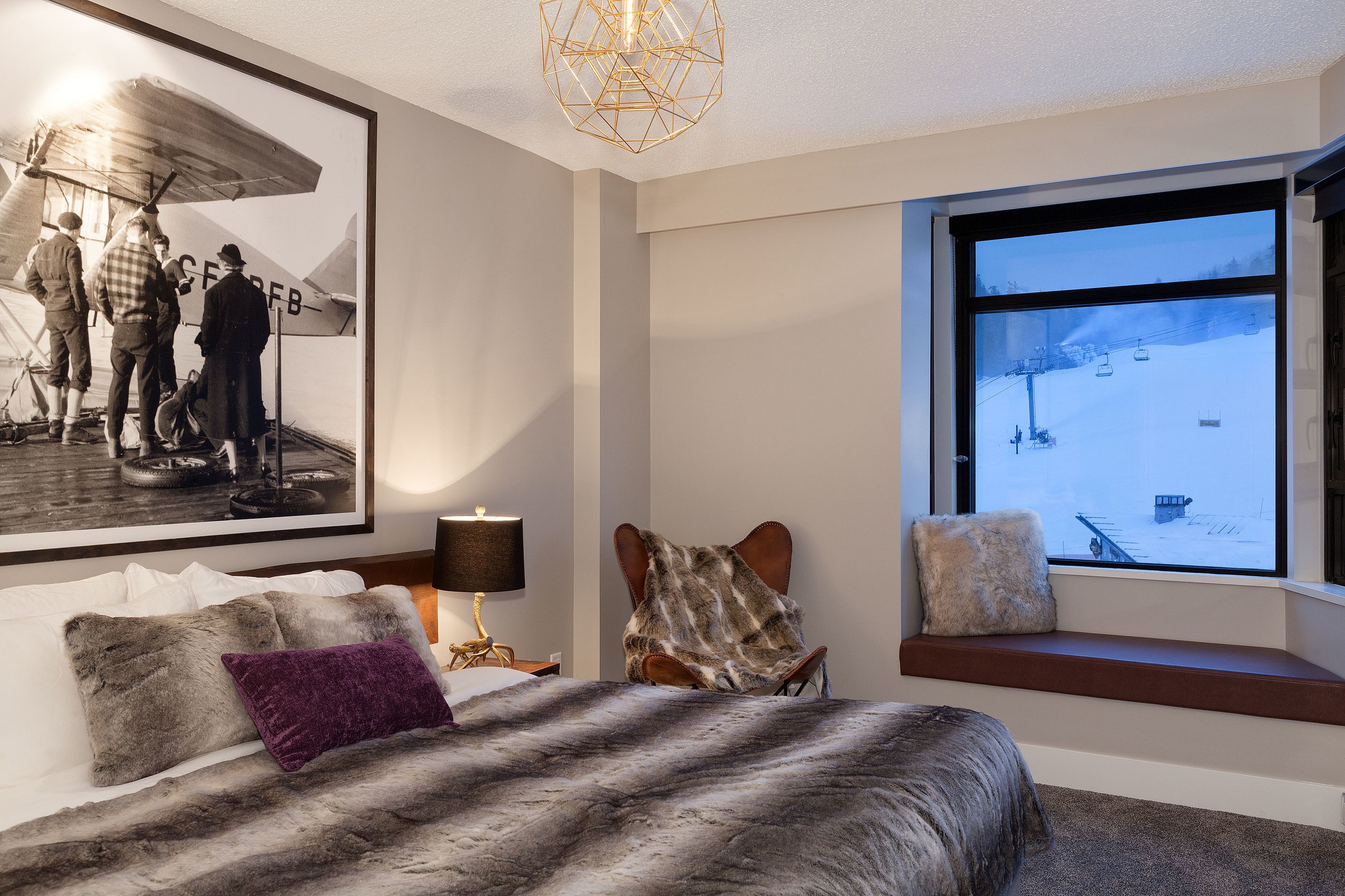 Un dormitorio con un ventanal grande, una cama con una manta de piel sintética, una silla con un cojín de piel sintética y un cuadro con una fotografía de un avión antiguo.