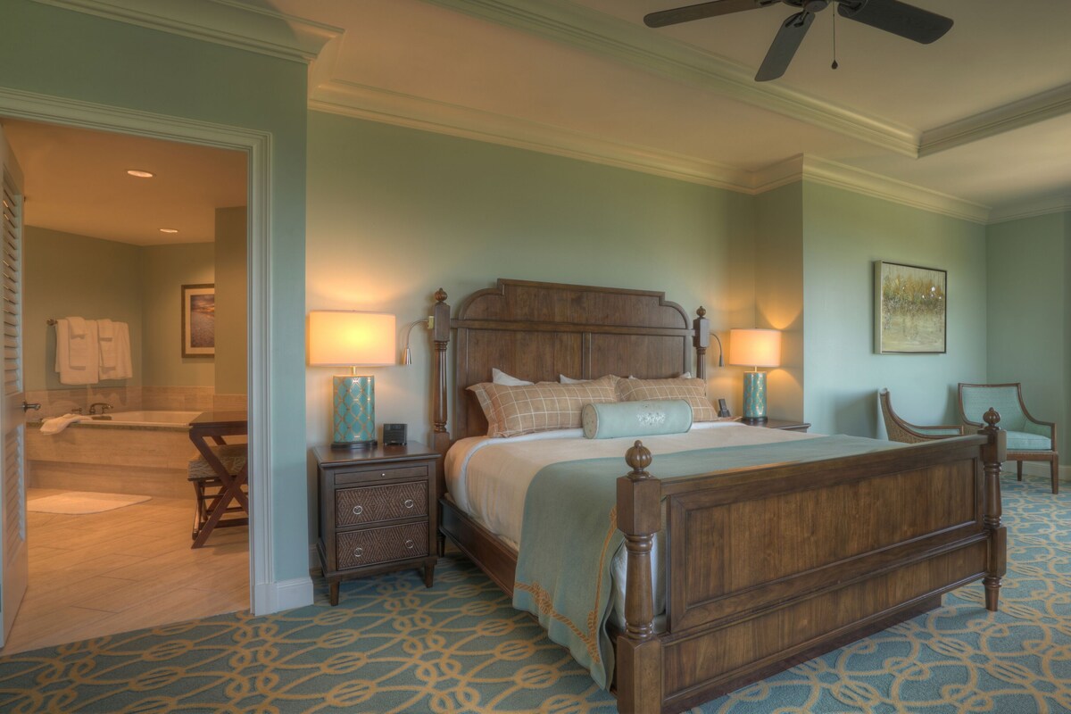 A bedroom with a large bed, two bedside tables, a chair, a desk, and a ceiling fan.