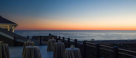 A terrace with tables and chairs overlooking the ocean at sunset.