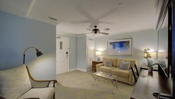 Un salon avec un canapé beige, une table basse en verre, une table d’appoint en bois, un ventilateur de plafond et un tableau accroché au mur.