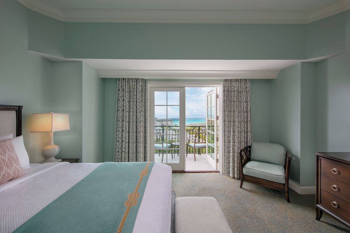 Habitación de hotel con cama, una silla, tocador y balcón con vistas.