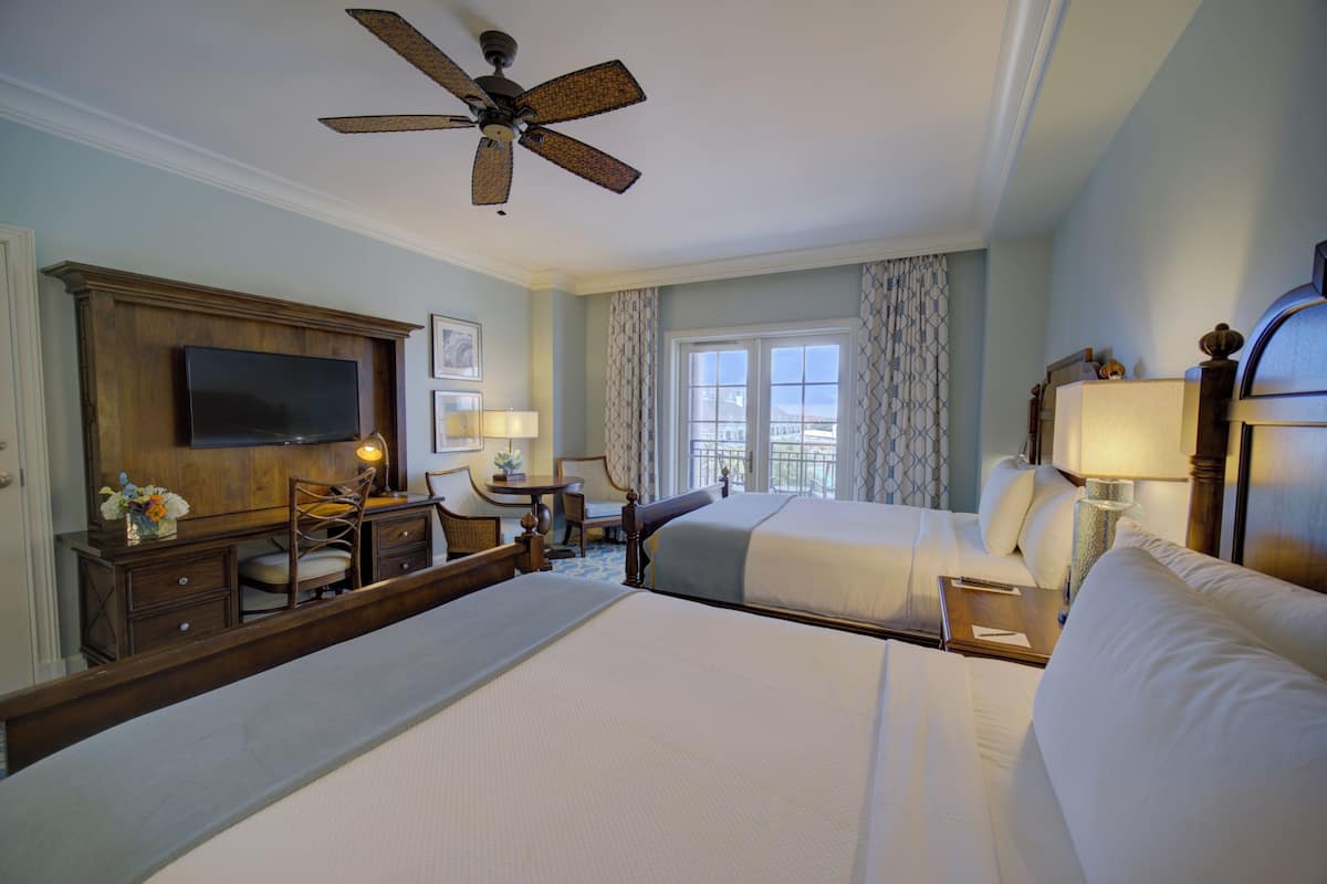 A hotel room with two beds, a desk, a chair, a television, and a ceiling fan.