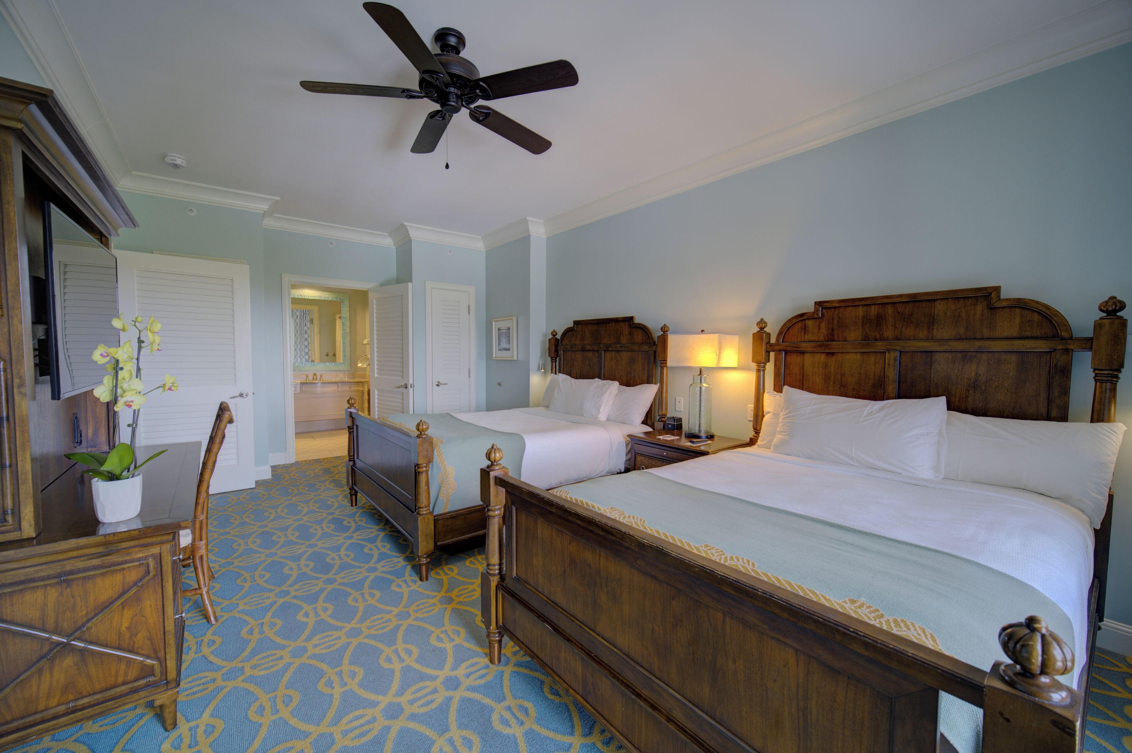 A hotel room with two beds, a ceiling fan, a wooden headboard, a nightstand, a lamp, a dresser, and a potted plant.