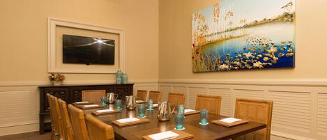 A dining room with a long table, chairs, a television, and a large painting on the wall.