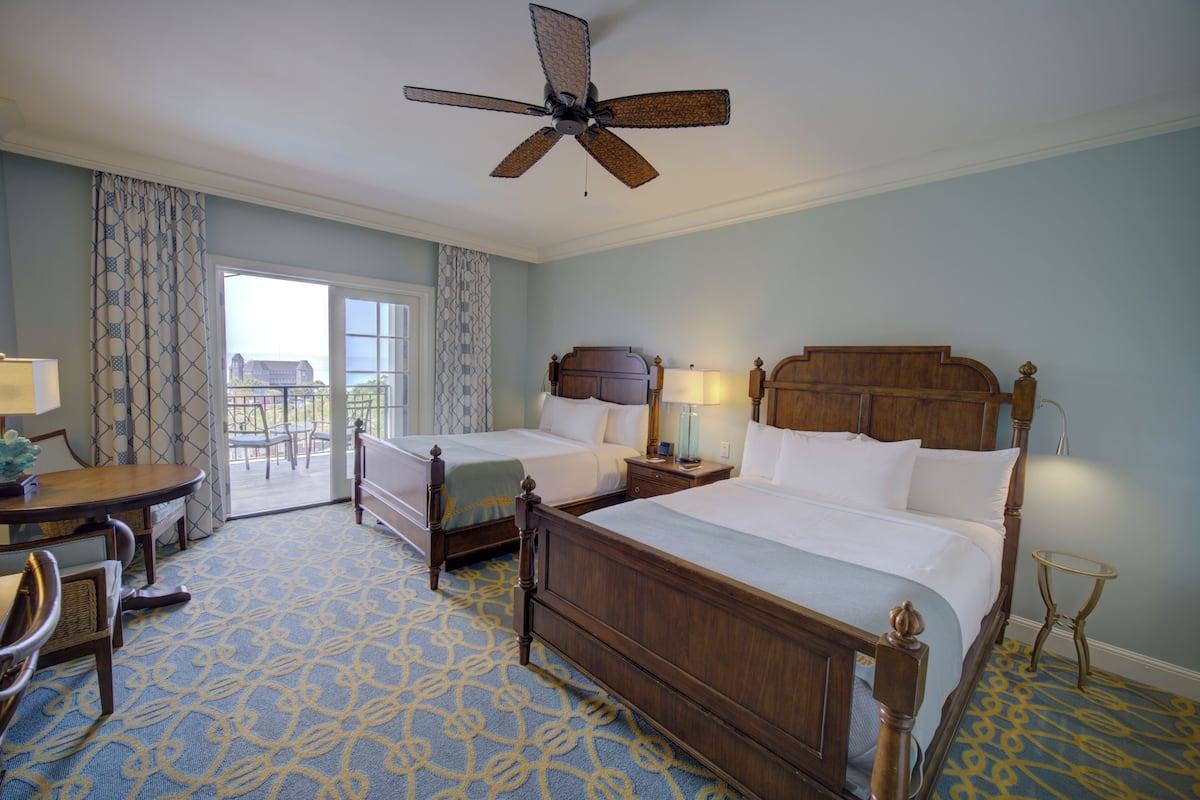 A hotel room with two beds, a dining table, chairs, a ceiling fan, and a view of the outdoors.