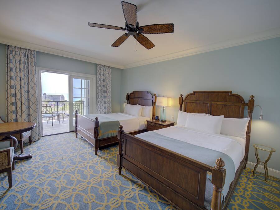 A hotel room with two beds, a dining table, chairs, a ceiling fan, and a view of the outdoors.
