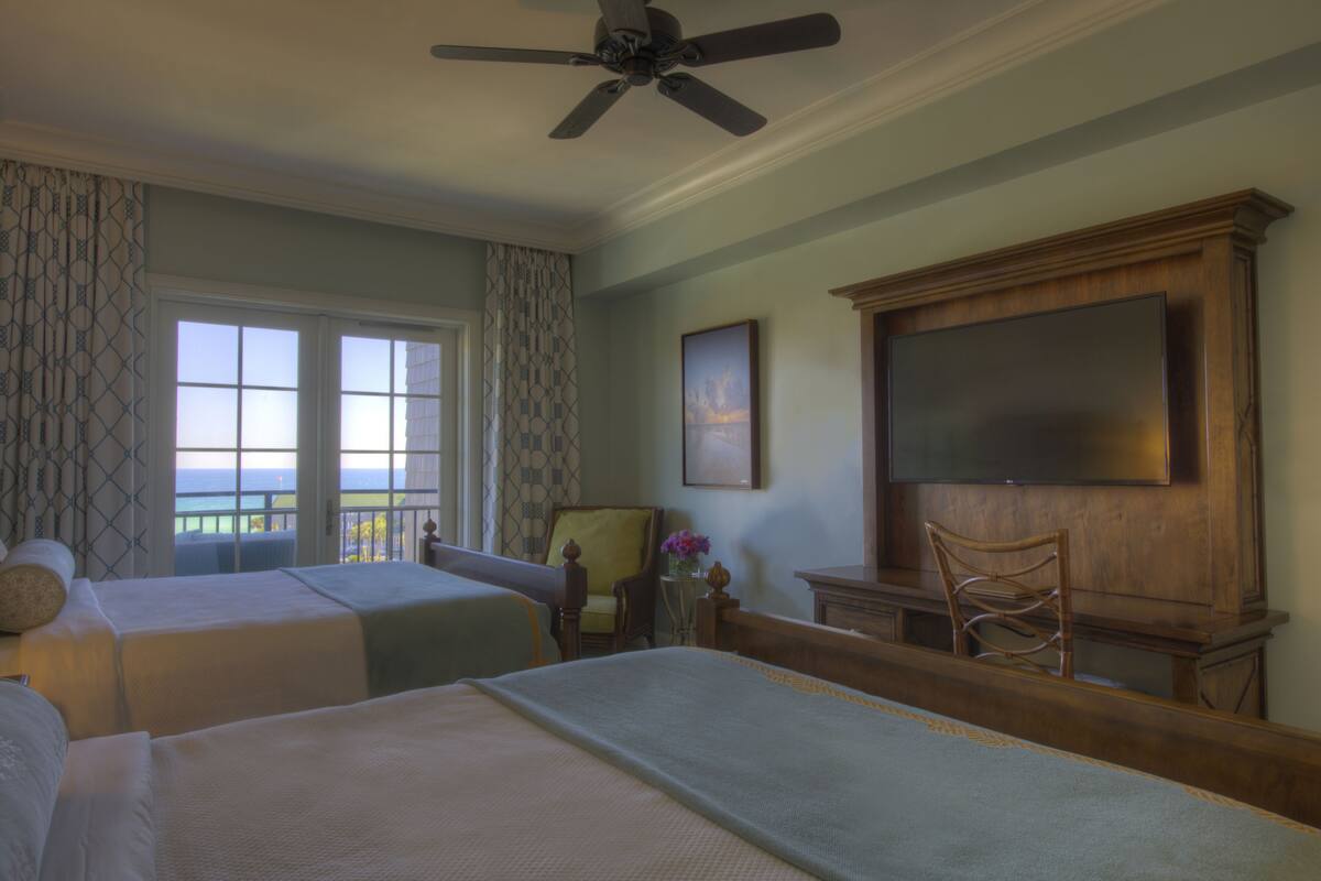 Habitación de hotel con dos camas, ventilador de techo, televisión, una silla y vista al océano.