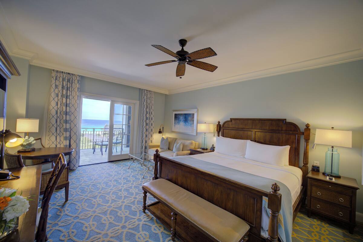 A hotel room with a large bed, a desk with a lamp, a bench, a nightstand, and a ceiling fan.