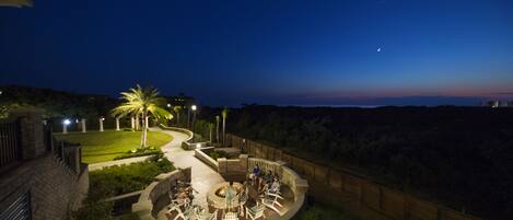 A nighttime outdoor patio area with seating and a fire pit, surrounded by greenery and a view of the city skyline.