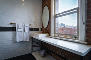 Un bagno con piastrelle bianche tipo metro, un lavandino bianco, uno specchio e una finestra con vista sugli edifici.