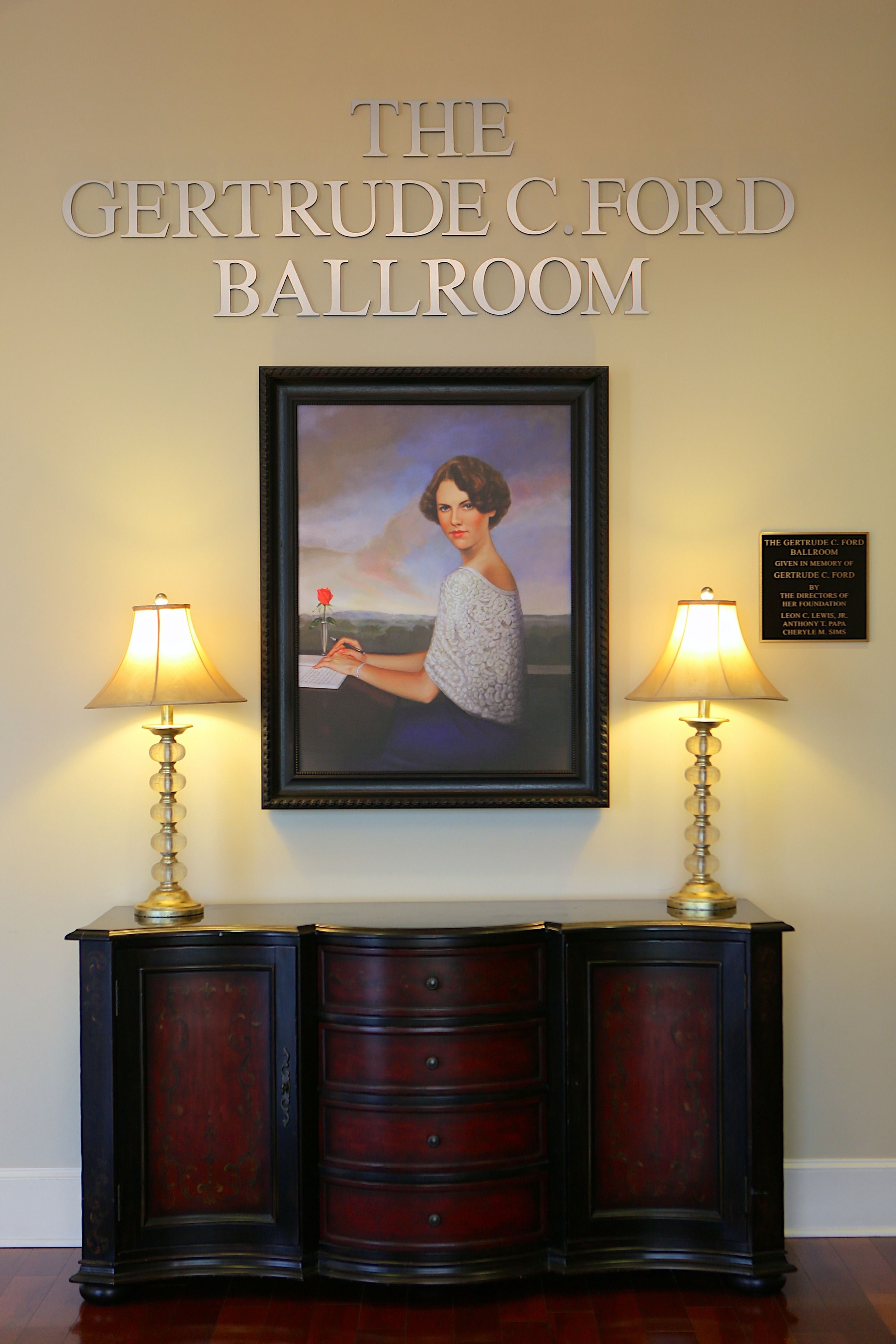 A framed portrait with a lamp on either side, a dark wooden cabinet below, and a plaque above the portrait.
