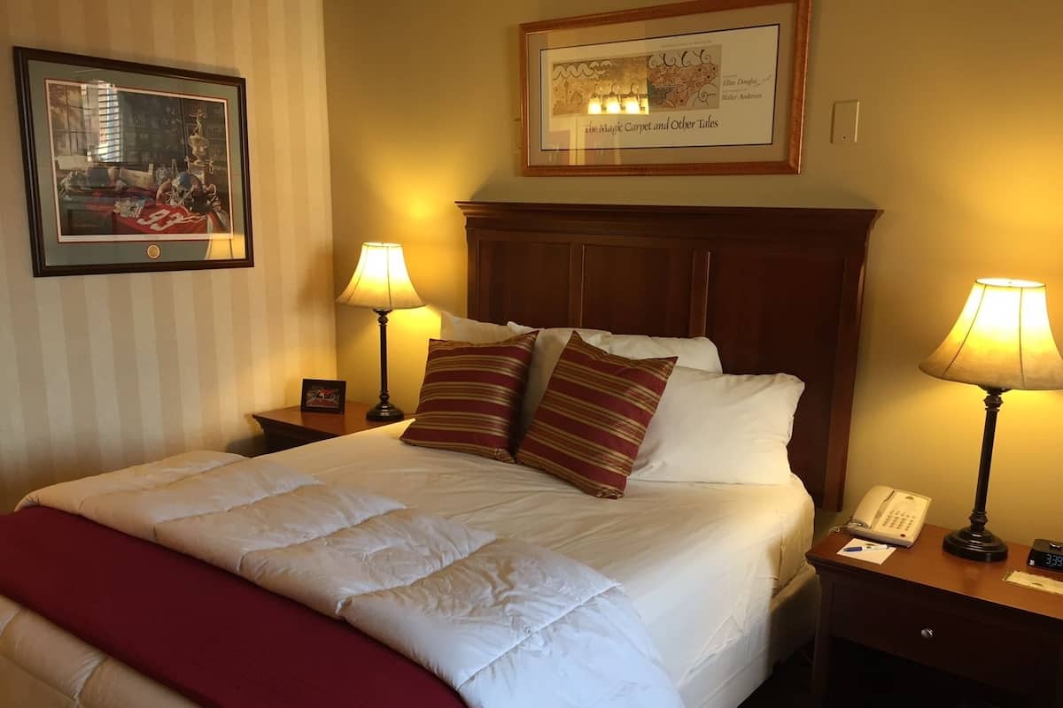 A neatly made bed with a red blanket, two bedside lamps, a framed picture above the bed, and a wooden nightstand with a phone and a clock.