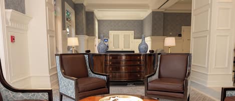 A lobby with a central wooden table, two brown leather chairs, a blue patterned sofa, and a decorative vase.