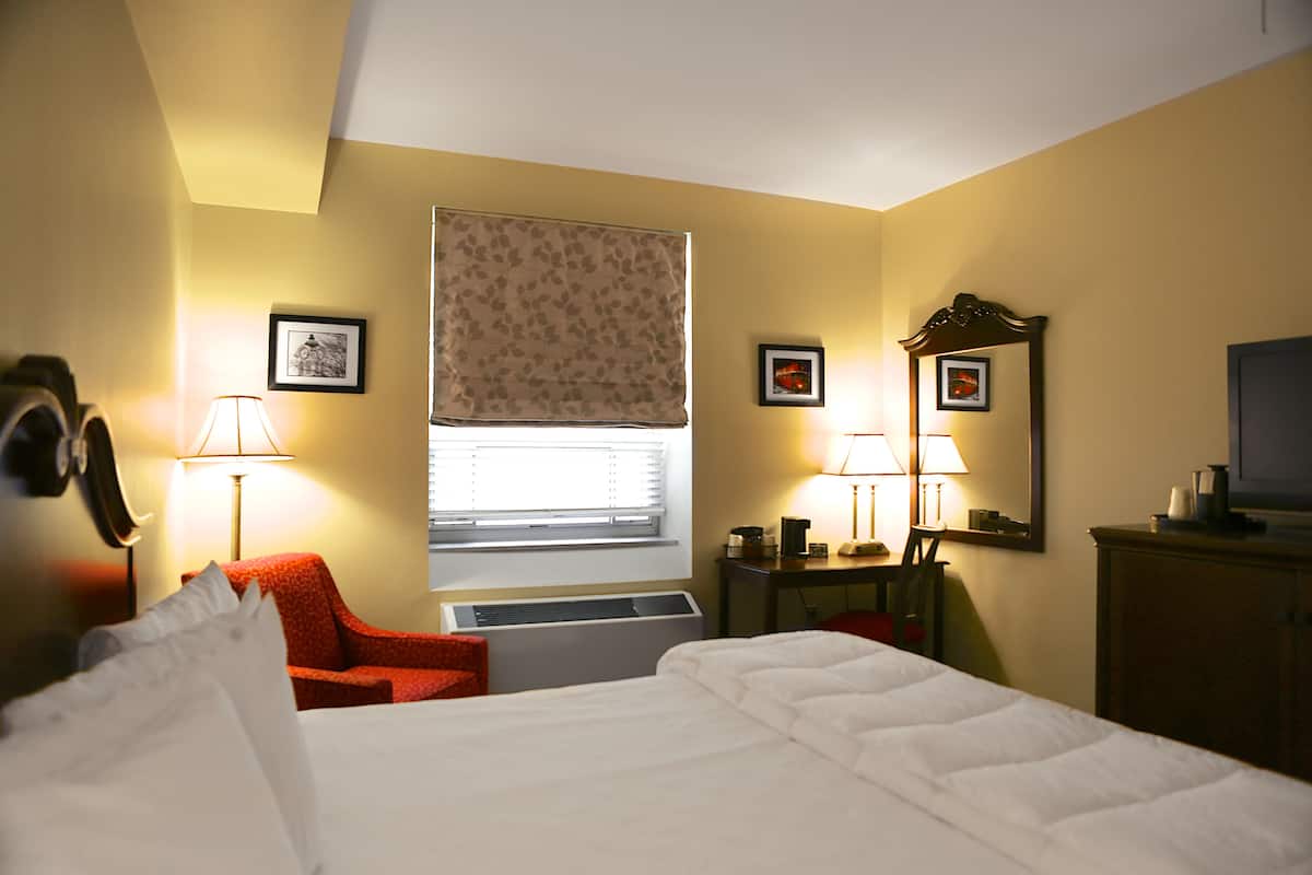A hotel room with a bed, a red chair, a desk with a lamp, and a window with a leaf-patterned blind.