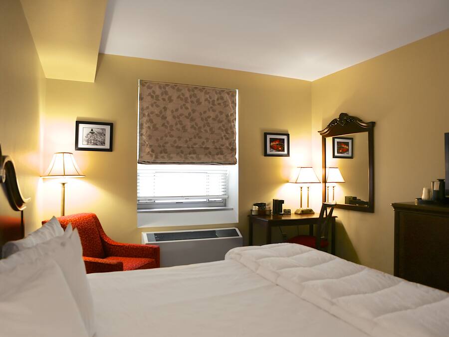 Una habitación de hotel con una cama, una silla roja, un escritorio con lámpara y una ventana con persiana de hojas.