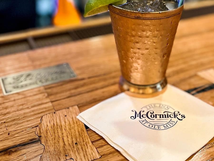 A tall glass with a lime wedge on a wooden table, a napkin with a logo, and a blurred background.