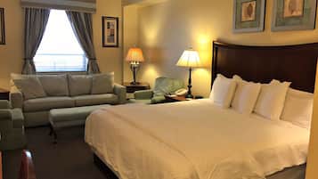 A hotel room with a large bed, a sofa, a chair, a nightstand with a lamp, and a window with curtains.