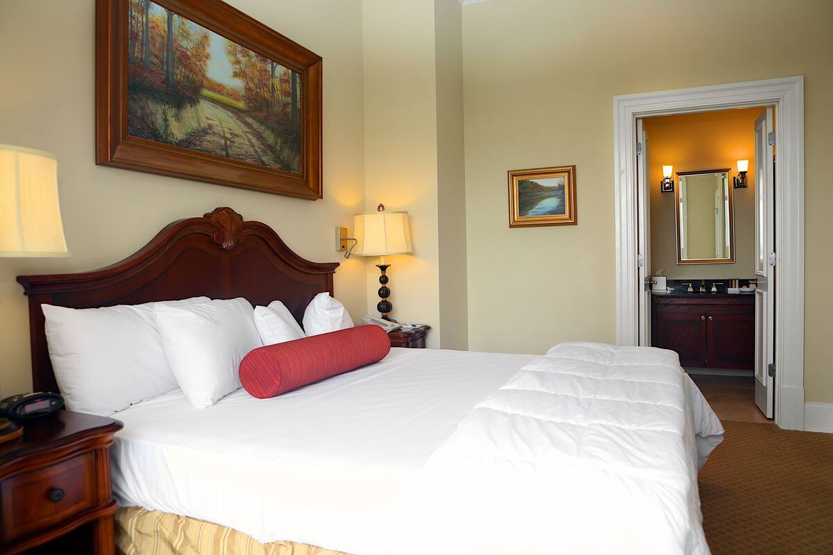 A hotel room with a large bed, a nightstand, a lamp, a painting, a mirror, and a door leading to a bathroom.