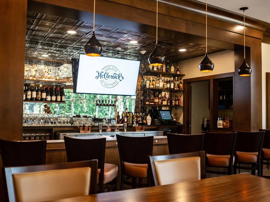 A bar area with a large screen displaying "The Inn McCormick's Aloe Mint" logo, wooden bar counter with bottles, and seating area with tables and chairs.