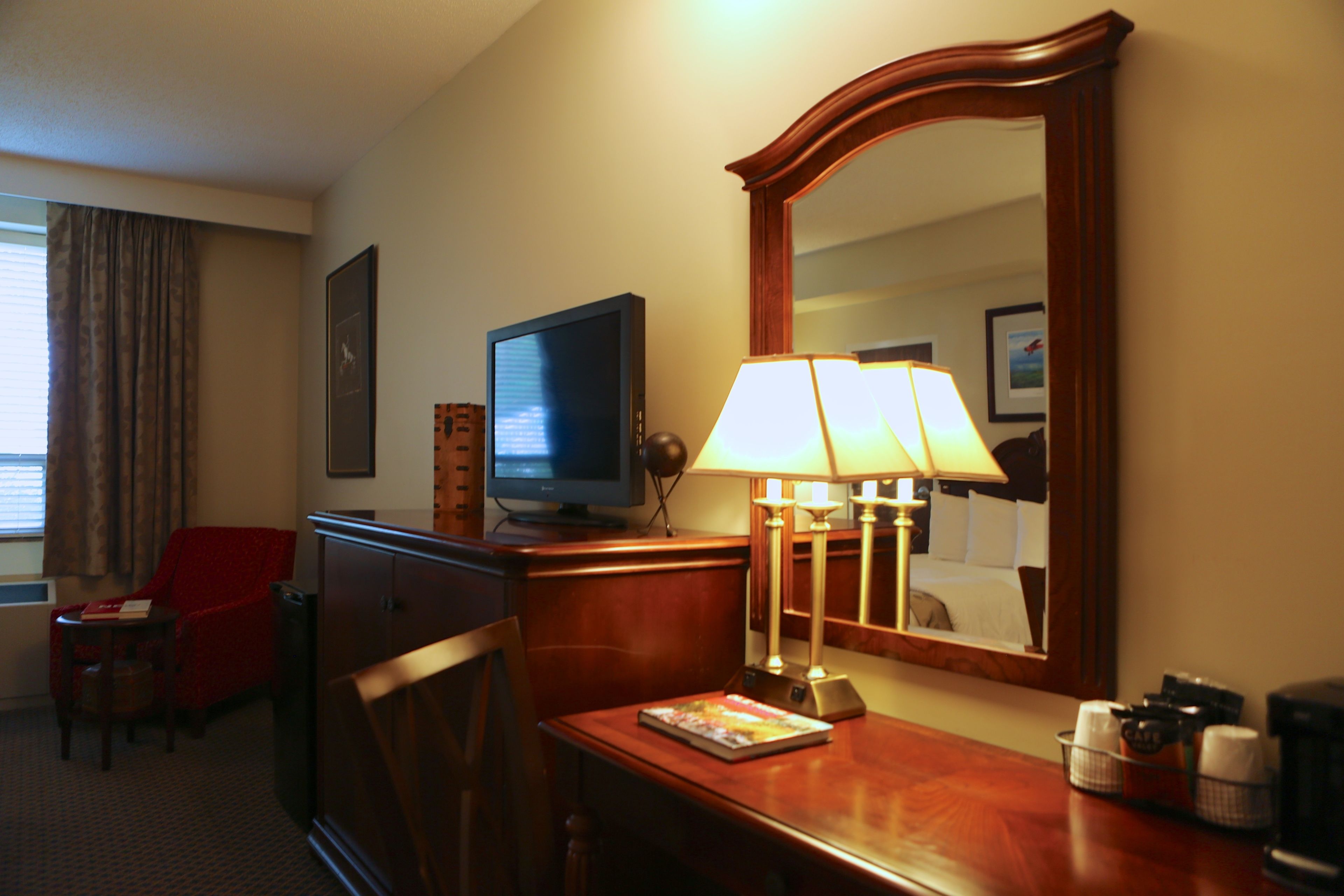 Ein Hotelzimmer mit Fernseher, Schreibtisch mit Lampe, Stuhl und Spiegel.