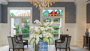 A lobby with a large, ornate chandelier, a round table with a vase of flowers, two patterned chairs, and a view through a stained glass window. - The Inn at Ole Miss (University)