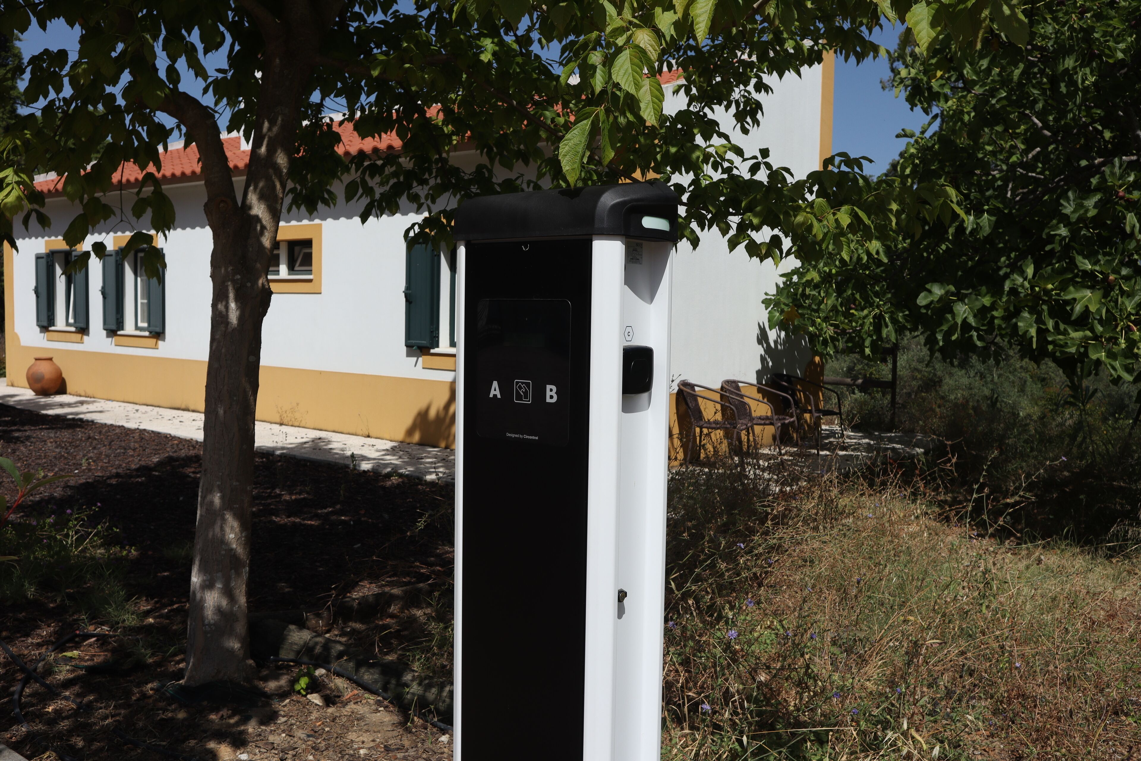 A black and white electrical box with symbols for a public charging station, located outdoors near a building with a yellow facade and a tree.
