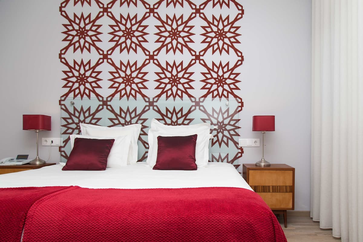 A neatly made bed with a red blanket, two red lamps on either side, and a patterned wall behind.