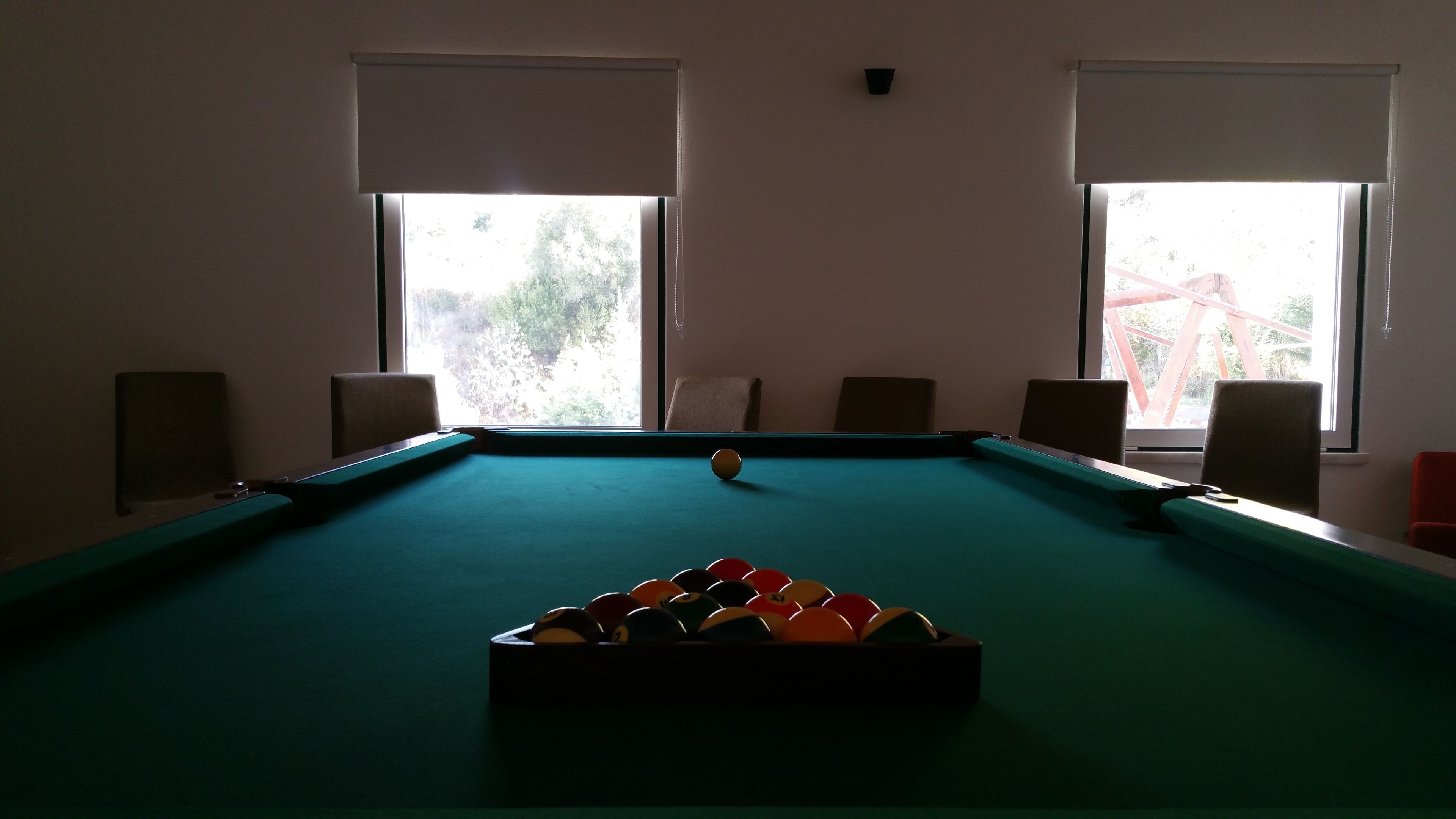 A pool table with a set of balls in the center, two windows with blinds, and four chairs around the table.