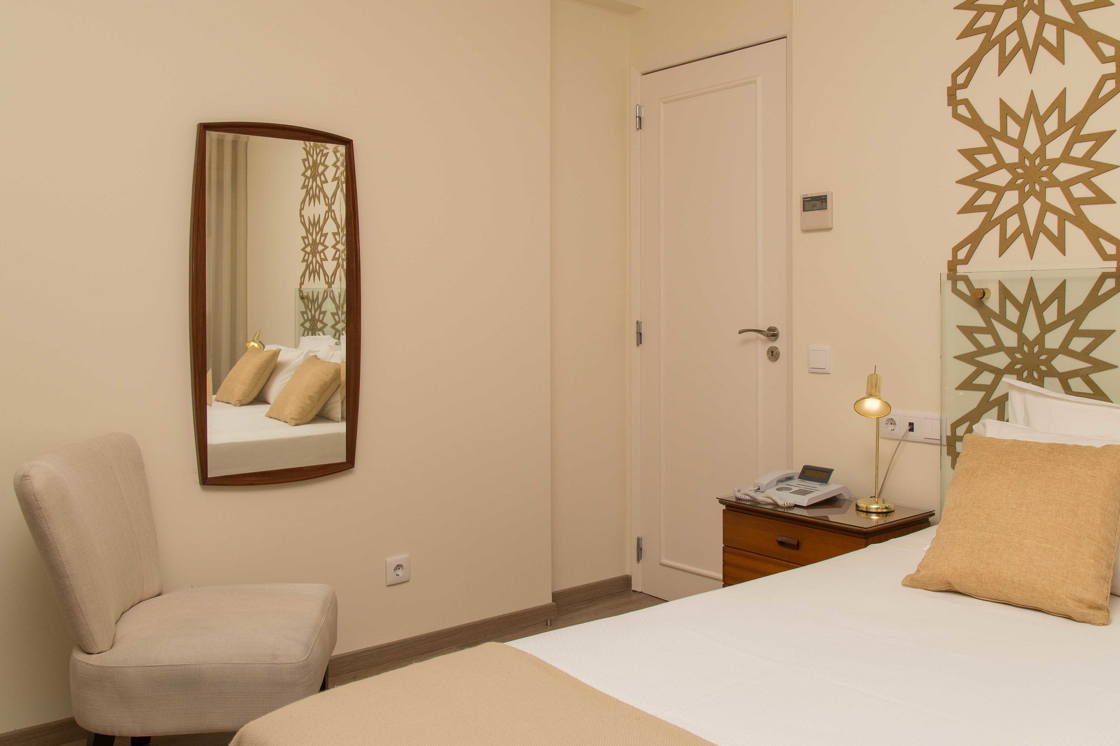 A hotel room with a bed, a chair, a bedside table with a lamp, and a mirror on the wall.