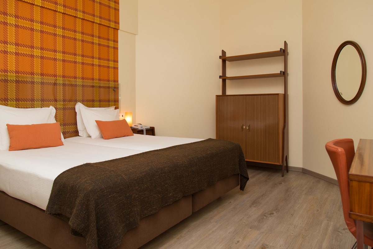 A hotel room with a bed, a wooden wardrobe, a mirror, and a small table with a chair.