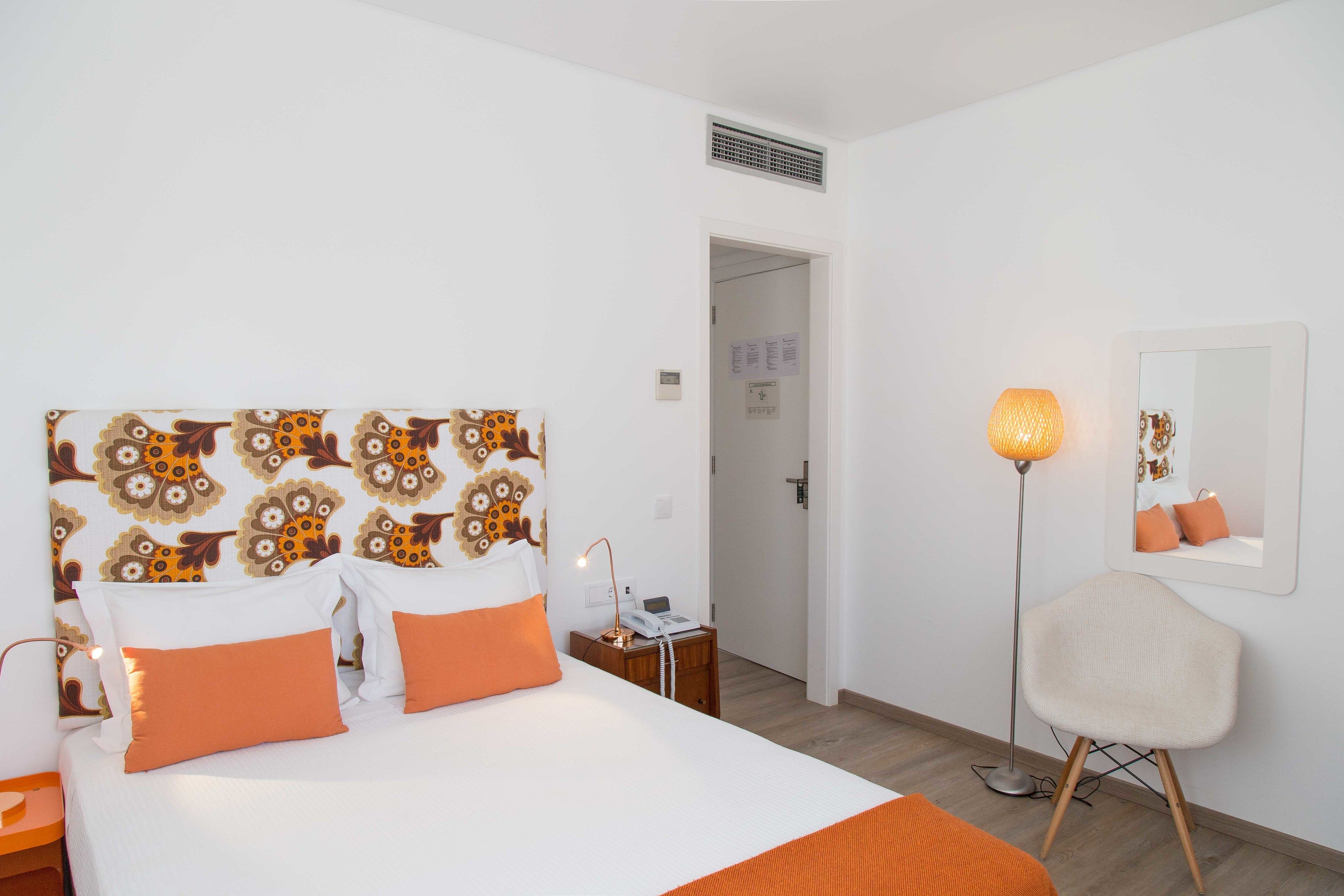 Habitación de hotel con cabecera estampada, mesita de noche con lámpara, una silla y un espejo en la pared.