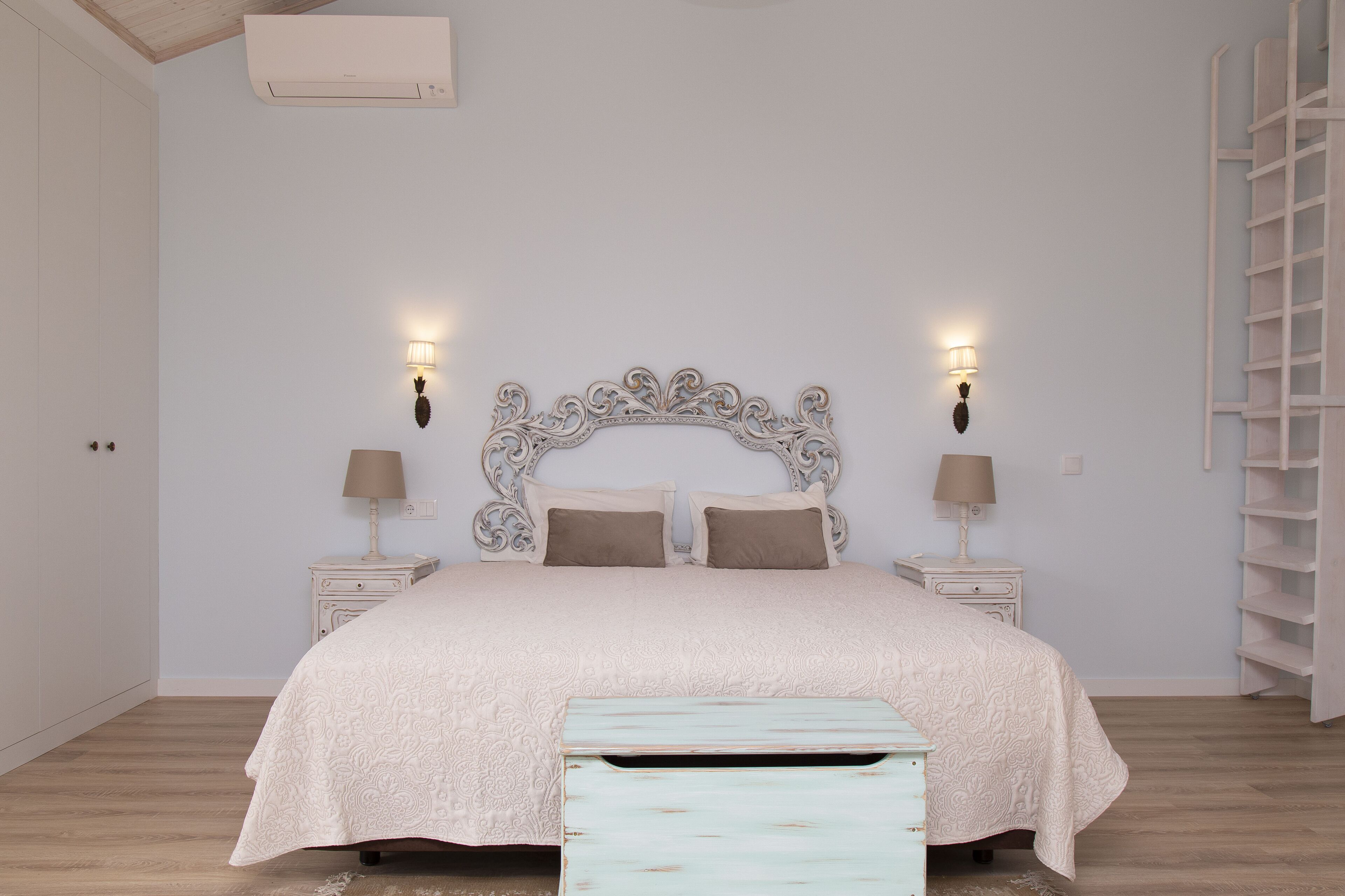 Une chambre à coucher avec un grand lit, deux tables de chevet avec des lampes, un banc en bois et une armoire blanche.