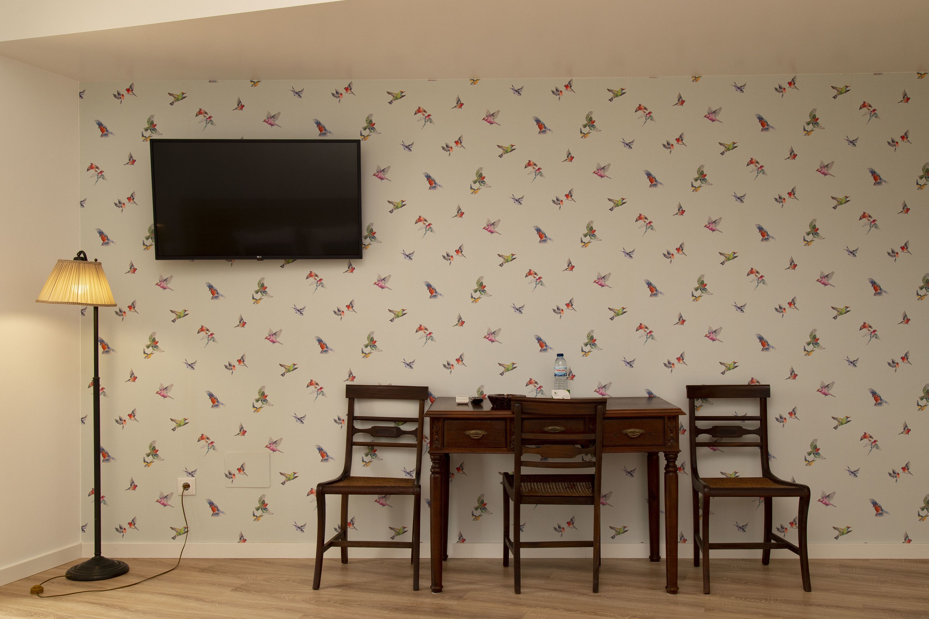 Una habitación con una televisión de pared, dos sillas de madera, un escritorio y una lámpara de pie.