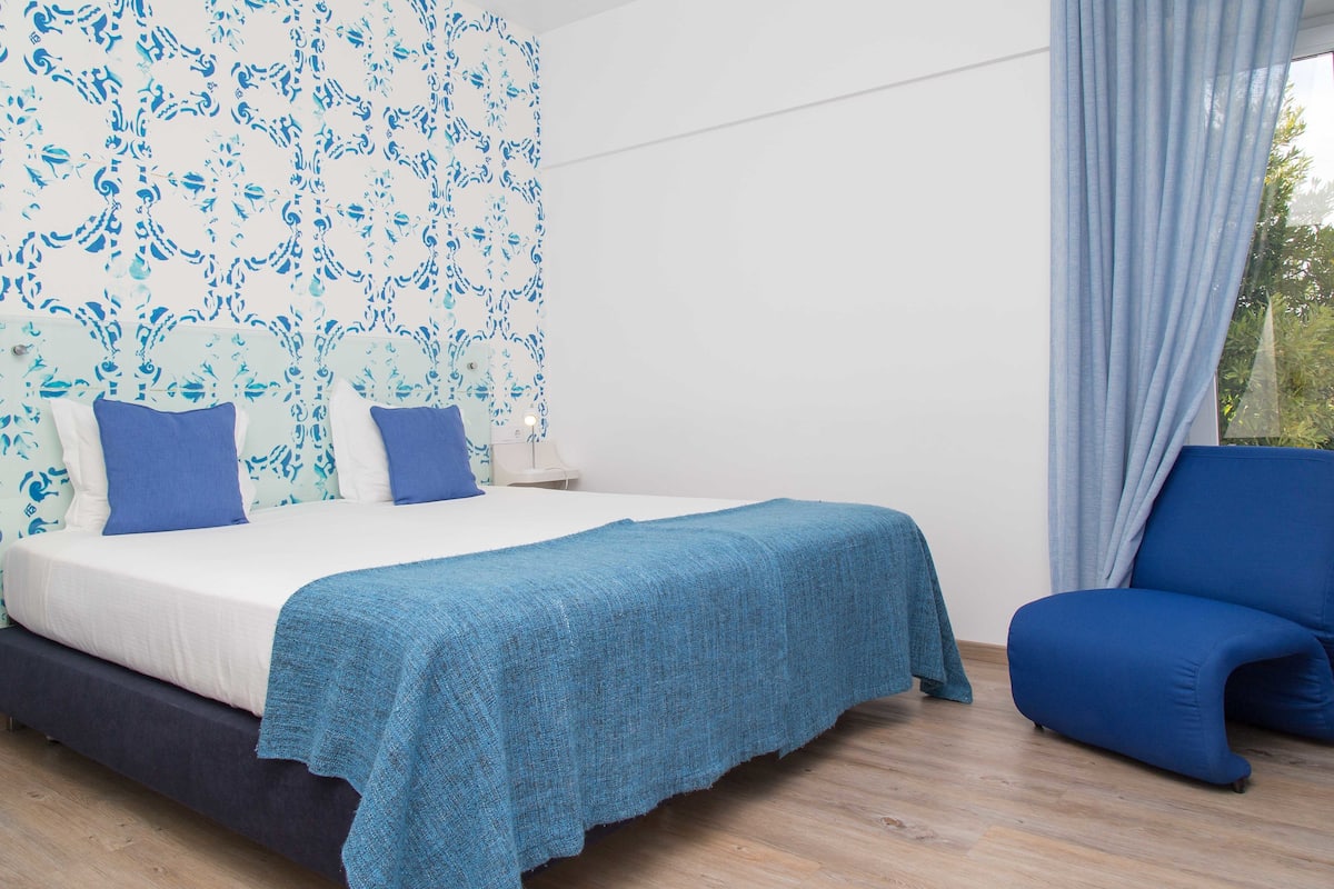A bedroom with a bed, blue pillows, a blue chair, and a window with curtains.