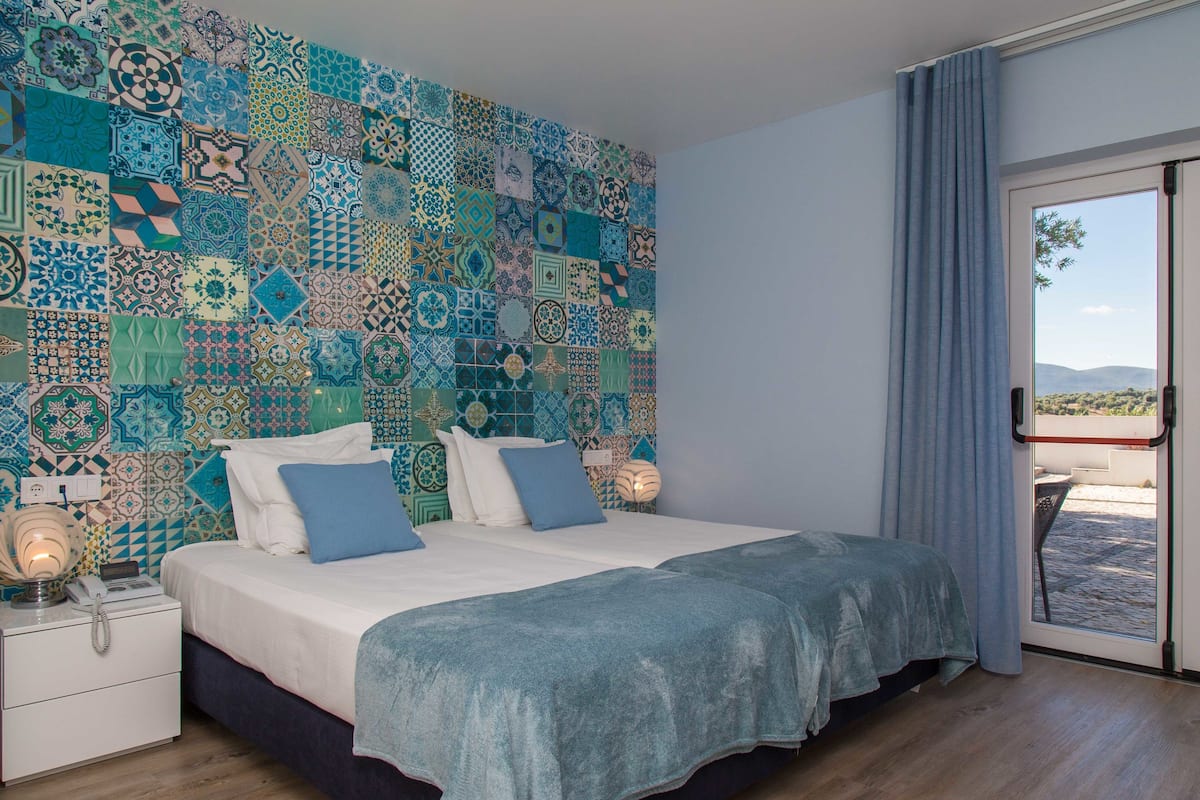 A bedroom with a patterned wall, a bed with blue bedding, a bedside table with a lamp, and a view through a glass door.