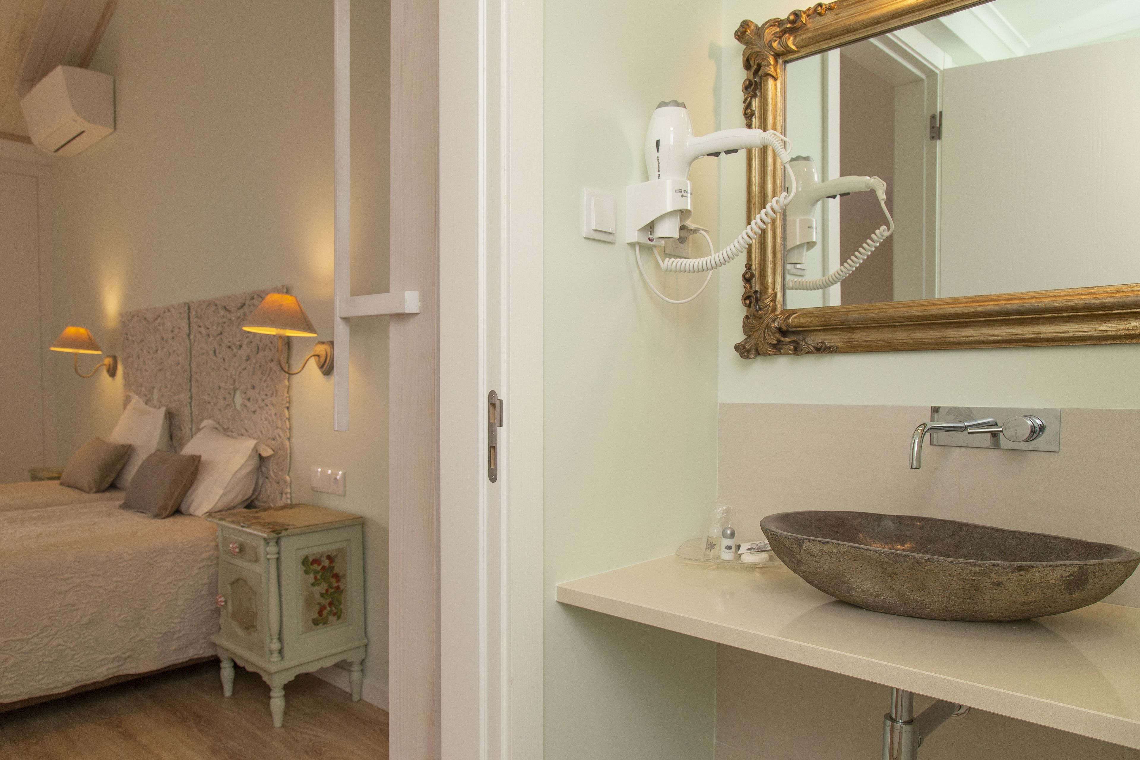 Un dormitorio con cama, lámparas de noche, mesita de noche, espejo y baño con lavabo y secador de pelo.