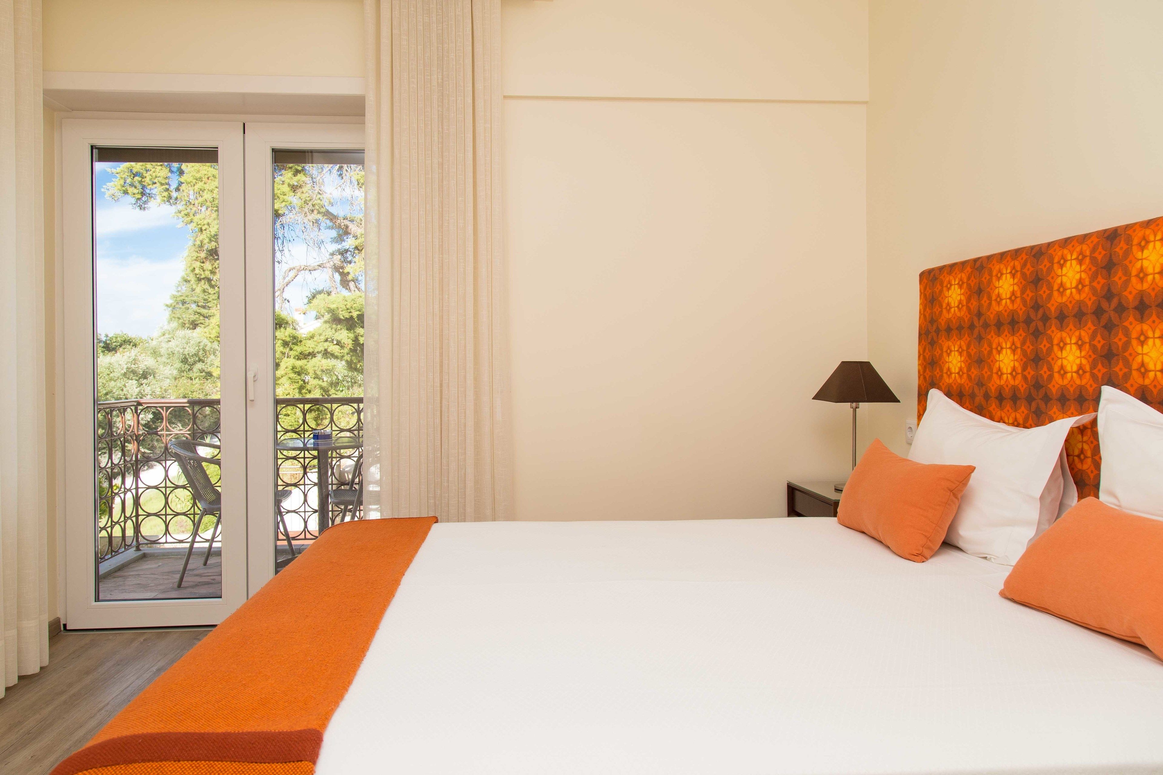 Habitación de hotel con una cama grande, ropa de cama naranja, una mesita de noche con lámpara y un balcón con mobiliario exterior.