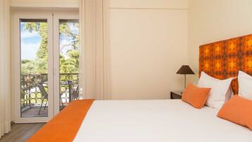Habitación de hotel con una cama grande, ropa de cama naranja, una mesita de noche con lámpara y un balcón con mobiliario exterior.