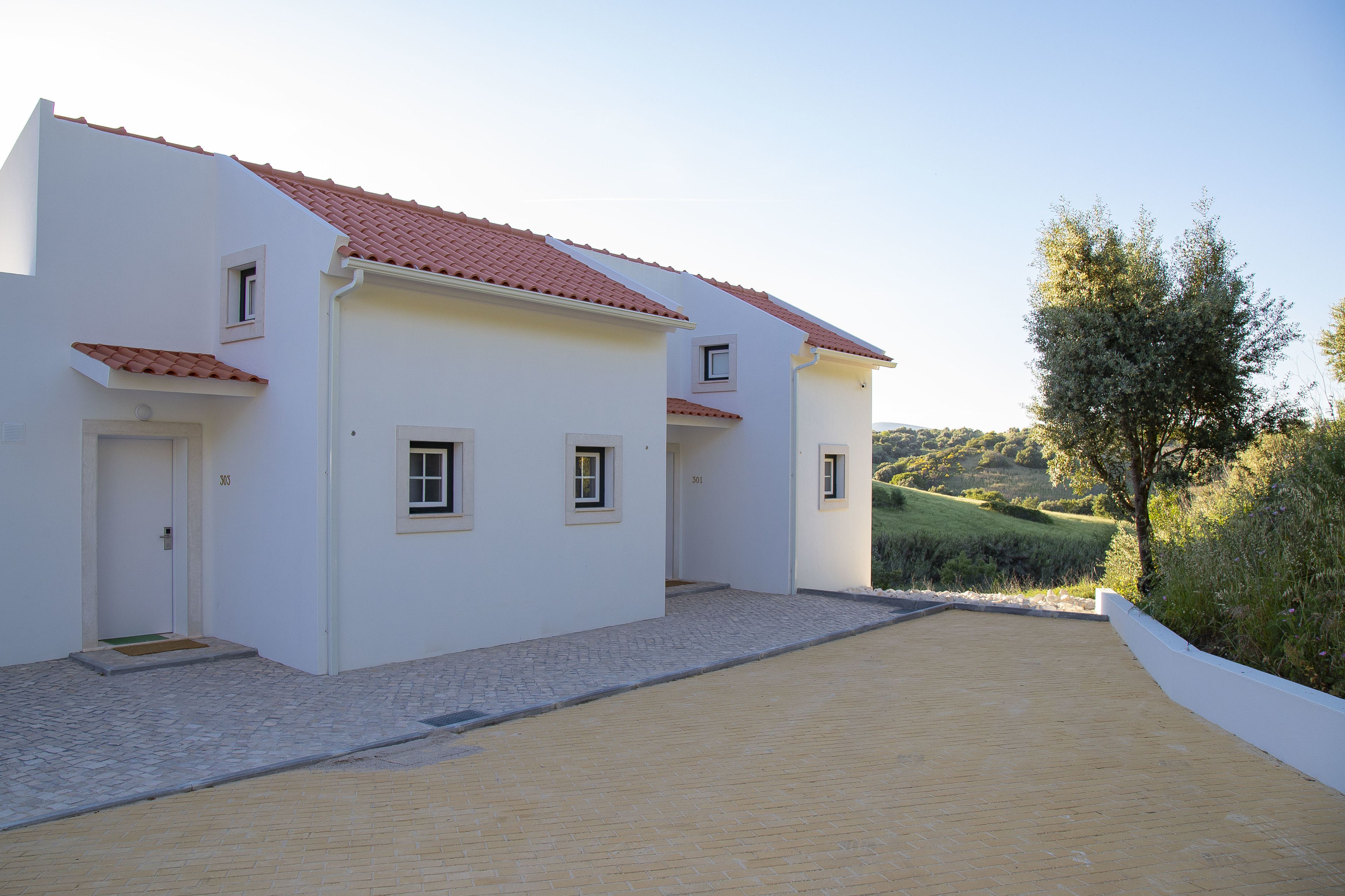 A two-story white building with a red-tiled roof, a paved driveway, and a view of a green hillside.