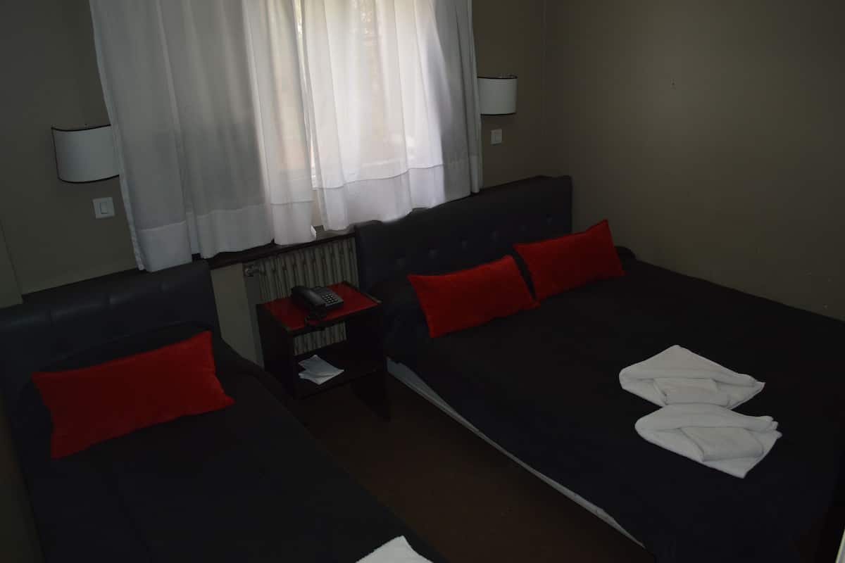 A hotel room with two beds, each with red and white pillows, a bedside table with a phone, and a window with sheer curtains.