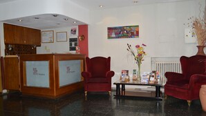 Ein Hotel-Empfangsbereich mit Holztresen, zwei roten Sesseln, einem Glastisch und einer Vase mit Blumen.