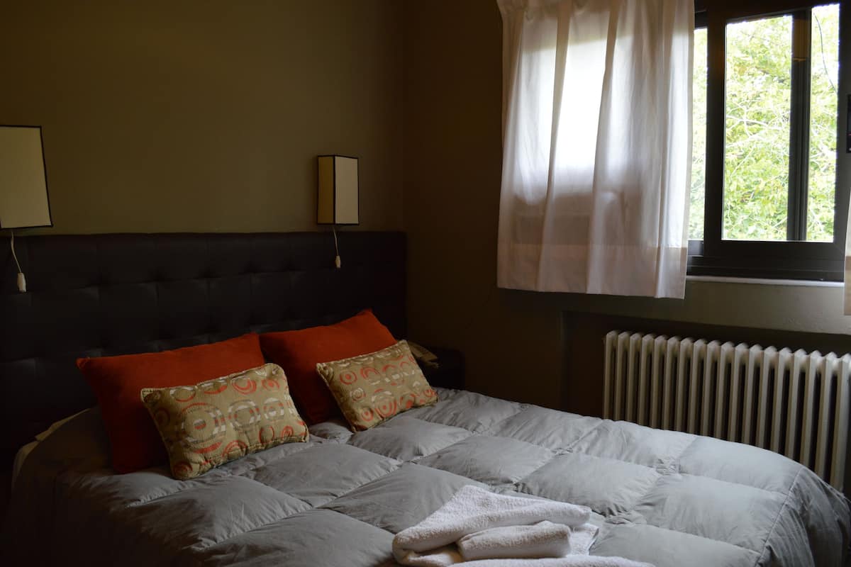 A neatly made bed with a grey duvet, two red pillows, and two patterned pillows. There is a window with sheer curtains and a radiator below it.