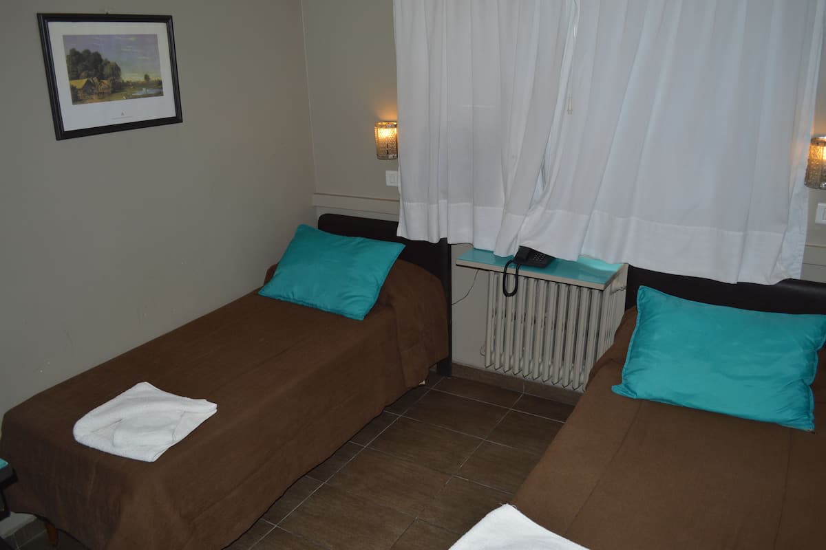 A room with two single beds, each with a teal pillow and a white towel. There is a framed picture on the wall, a radiator under the window, and white curtains.